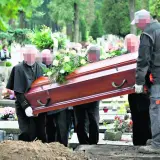 Pogrzeb na cmentarzu. Kilku mężczyzn niesie drewnianą trumnę ozdobioną kwiatami w kierunku grobu głębinowego.