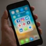 Dłoń trzyma smartfon z ikonami mediów społecznościowych. Folder "Social Networks" zawiera Facebooka, Instagram, Twitter, Google+, Pinterest, Tumblr, LinkedIn, WhatsApp i Messenger.