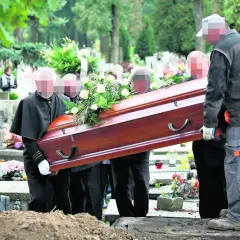 Pogrzeb na cmentarzu. Kilku mężczyzn niesie drewnianą trumnę ozdobioną kwiatami w kierunku grobu głębinowego.