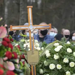 Drewniany krzyż z figurą Chrystusa na pogrzebie. Wieniec białych róż i czerwonych kwiatów. Ludzie w maseczkach. Co zrobić z krzyżem po pogrzebie?