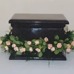 Czarna, kamienna urna ozdobiona girlandą z delikatnych, różowych kwiatów i zielonych liści. Kwiaty na trumnę symbolizują pożegnanie.