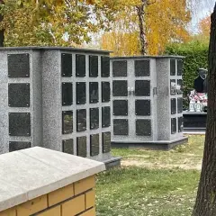 Nowoczesne kolumbarium rodzinne z szarego granitu, z wieloma pustymi tabliczkami na epitafia, otoczone jesienną zielenią.