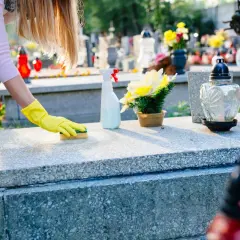 Kobieta w żółtych rękawiczkach czyści nagrobek, pokazując jak usunąć wosk z pomnika. Na stole leży butelka ze sprayem i kwiaty.
