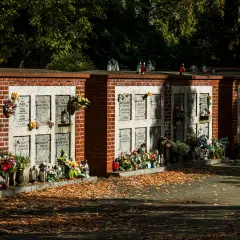 Cmentarne kolumbarium z rzędami nisz na urny, ozdobione kwiatami i zniczami. Jesienne liście pokrywają ścieżkę.