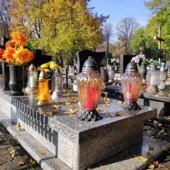 Jesienny dzień na cmentarzu. Na kamiennym, murowanym grobie palą się znicze, a obok stoją świeże kwiaty.