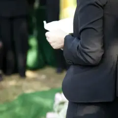 Kobieta w czerni trzyma kartkę, przygotowując się do mowy pogrzebowej. W tle widać żałobników.