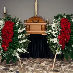 Dwa wieńce pogrzebowe z czerwonych róż i białych kwiatów stoją obok drewnianej trumny. Czy na kremację kupuje się kwiaty? Tak, są one wyrazem pamięci.
