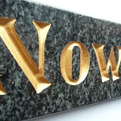 Złote litery kute na pomnik, wyraz "Nowa", na ciemnym, granitowym tle.