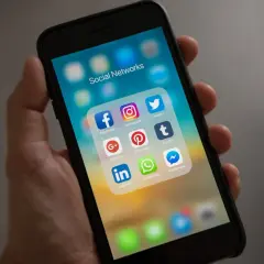 Dłoń trzyma smartfon z ikonami mediów społecznościowych. Folder "Social Networks" zawiera Facebooka, Instagram, Twitter, Google+, Pinterest, Tumblr, LinkedIn, WhatsApp i Messenger.