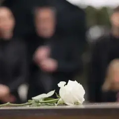 Biała róża na kamiennej płycie, w tle zamazane sylwetki żałobników. Czas organizacji pogrzebu jest trudny.