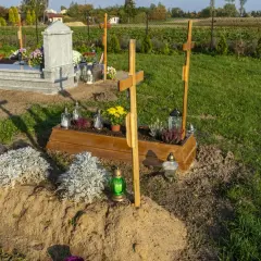 Drewniana trumna z kwiatami i krzyżami, obok kamienny nagrobek. Tak wygląda grób, ozdobiony zniczami i zielenią.
