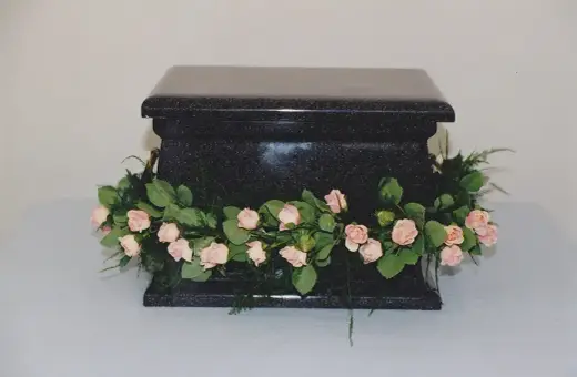Czarna, kamienna urna ozdobiona girlandą z delikatnych, różowych kwiatów i zielonych liści. Kwiaty na trumnę symbolizują pożegnanie.