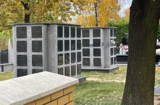 Nowoczesne kolumbarium rodzinne z szarego granitu, z wieloma pustymi tabliczkami na epitafia, otoczone jesienną zielenią.
