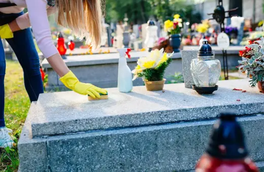 Kobieta w żółtych rękawiczkach czyści nagrobek, pokazując jak usunąć wosk z pomnika. Na stole leży butelka ze sprayem i kwiaty.