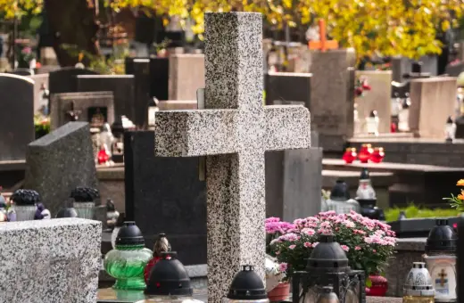 Kamienny krzyż i znicze na cmentarzu. W tle nagrobki i jesienne liście. Warto zadbać o porządek, by wiedzieć, jak usunąć chwasty na cmentarzu.