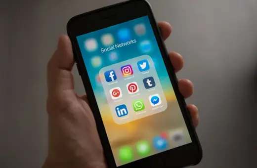 Dłoń trzyma smartfon z ikonami mediów społecznościowych. Folder "Social Networks" zawiera Facebooka, Instagram, Twitter, Google+, Pinterest, Tumblr, LinkedIn, WhatsApp i Messenger.