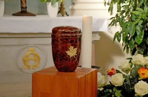 Elegancka urna z prochem, ozdobiona złotym liściem, stoi obok świecy i kwiatów.