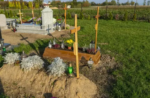 Drewniana trumna z kwiatami i krzyżami, obok kamienny nagrobek. Tak wygląda grób, ozdobiony zniczami i zielenią.