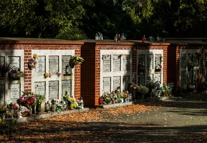 Cmentarne kolumbarium z rzędami nisz na urny, ozdobione kwiatami i zniczami. Jesienne liście pokrywają ścieżkę.