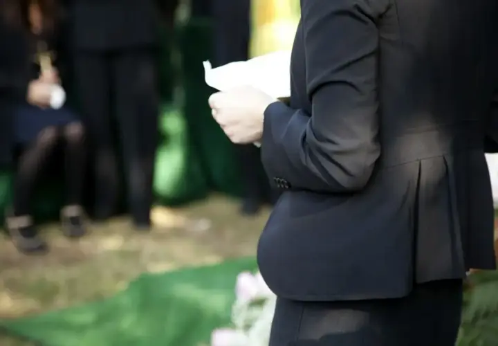 Kobieta w czerni trzyma kartkę, przygotowując się do mowy pogrzebowej. W tle widać żałobników.