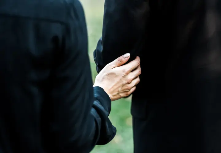 Dłoń obejmuje ramię, wspierając w trudnym momencie. To obraz żałoby i wspólnego przeżywania straty podczas orszaku pogrzebowego.