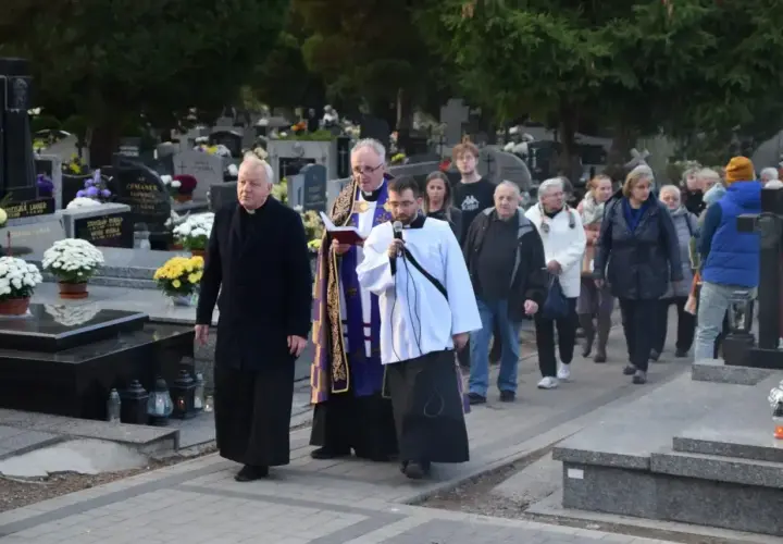 Ksiądz i ministrant niosą urnę na pogrzebie, a za nimi idzie żałobna procesja przez cmentarz.
