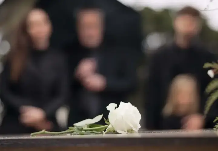Biała róża na kamiennej płycie, w tle zamazane sylwetki żałobników. Czas organizacji pogrzebu jest trudny.