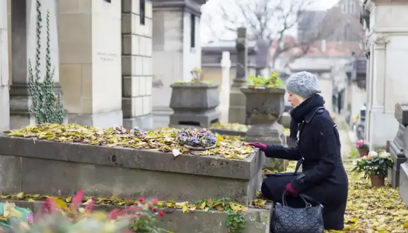 Kobieta klęczy przy grobie, na którym leżą jesienne liście. Przygotowuje się do przeniesienia urny z prochami na inny cmentarz.