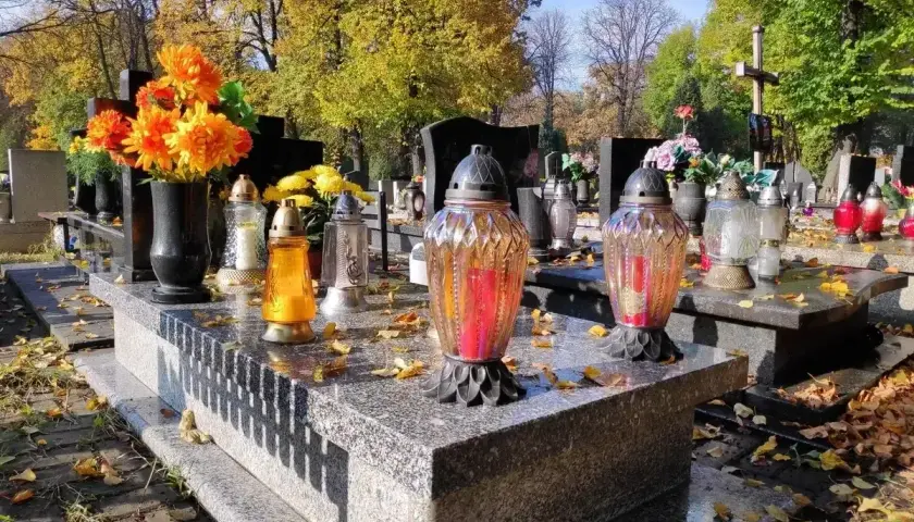 Jesienny dzień na cmentarzu. Na kamiennym, murowanym grobie palą się znicze, a obok stoją świeże kwiaty.