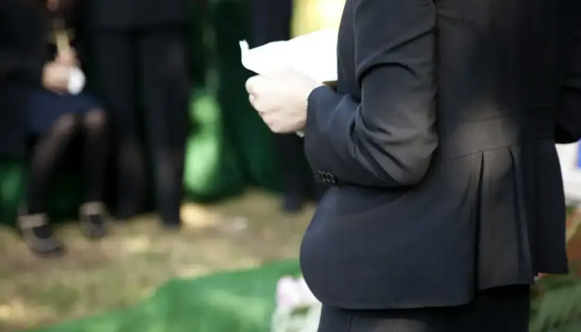 Kobieta w czerni trzyma kartkę, przygotowując się do mowy pogrzebowej. W tle widać żałobników.