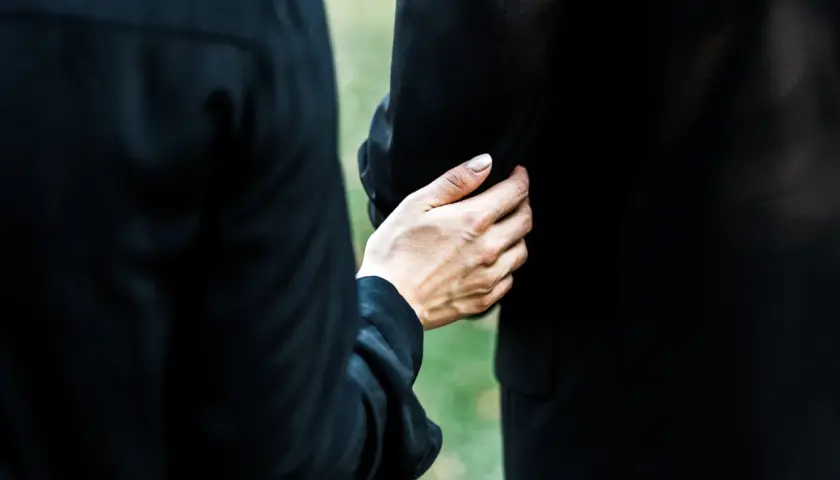 Dłoń obejmuje ramię, wspierając w trudnym momencie. To obraz żałoby i wspólnego przeżywania straty podczas orszaku pogrzebowego.
