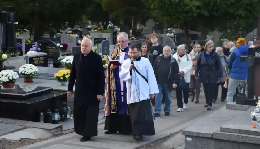 Ksiądz i ministrant niosą urnę na pogrzebie, a za nimi idzie żałobna procesja przez cmentarz.
