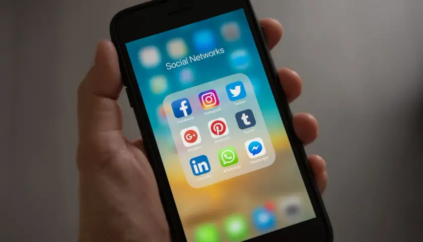 Dłoń trzyma smartfon z ikonami mediów społecznościowych. Folder "Social Networks" zawiera Facebooka, Instagram, Twitter, Google+, Pinterest, Tumblr, LinkedIn, WhatsApp i Messenger.