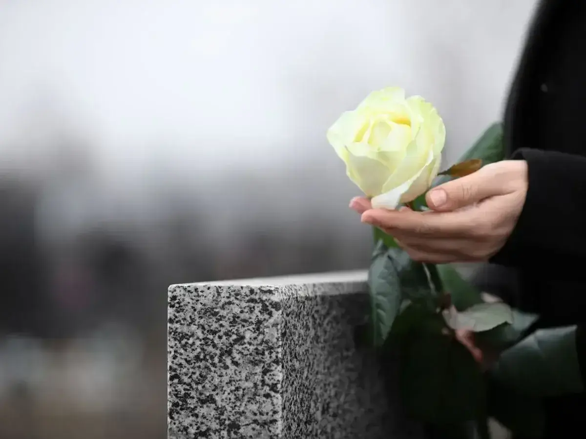 Dłoń trzyma białą różę przy nagrobku. Pytanie, jak długo nie można ruszać rzeczy zmarłego, pozostaje otwarte.