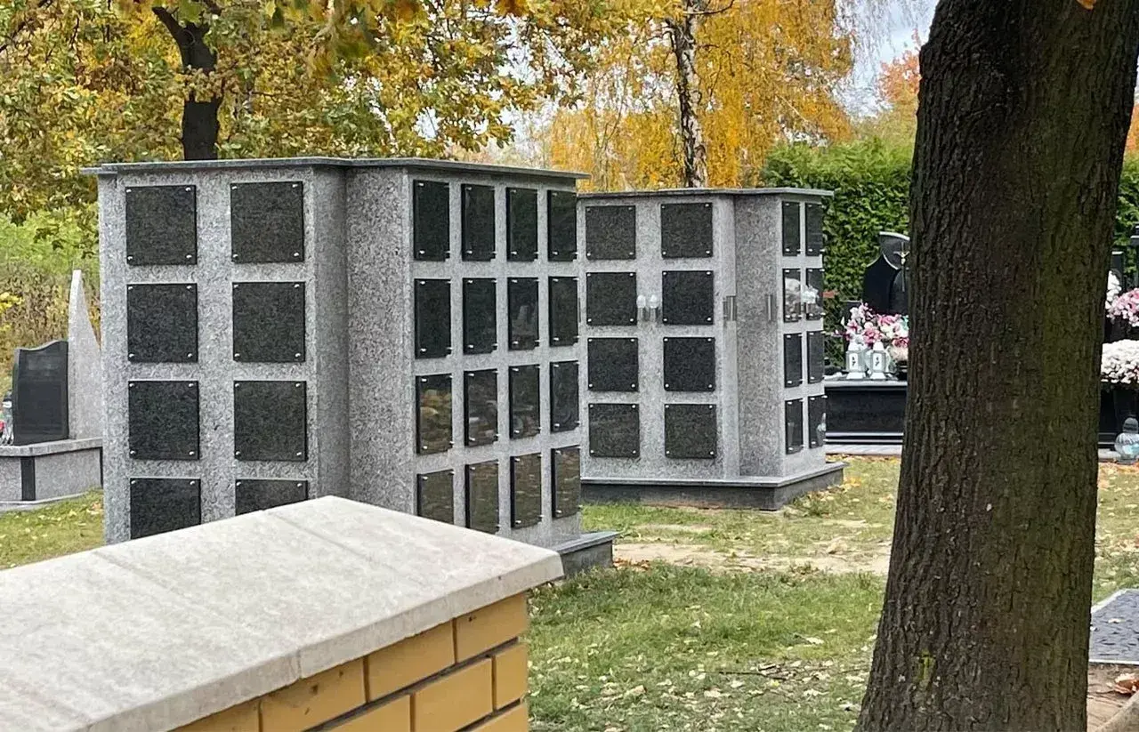 Nowoczesne kolumbarium rodzinne z szarego granitu, z wieloma pustymi tabliczkami na epitafia, otoczone jesienną zielenią.