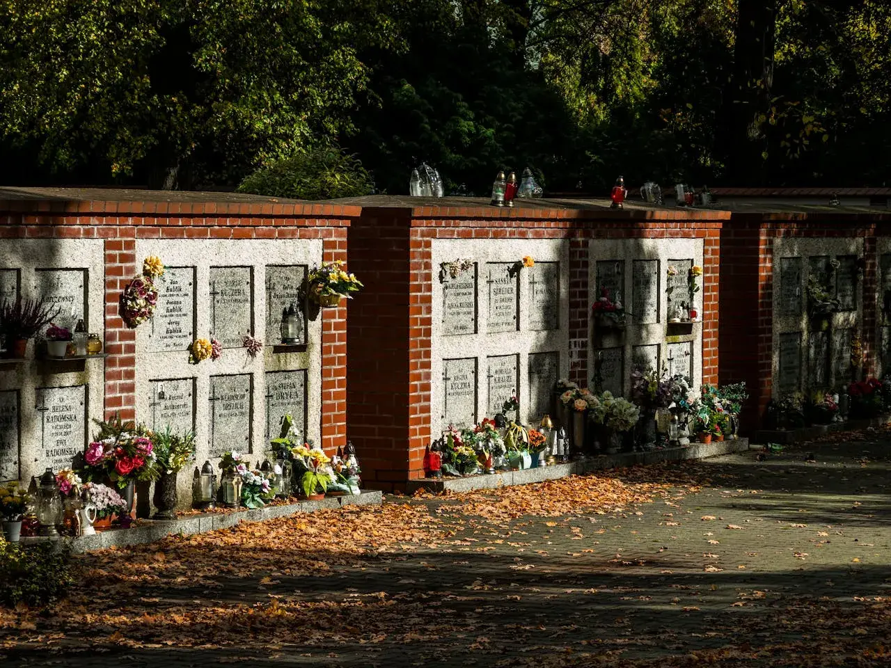 Cmentarne kolumbarium z rzędami nisz na urny, ozdobione kwiatami i zniczami. Jesienne liście pokrywają ścieżkę.