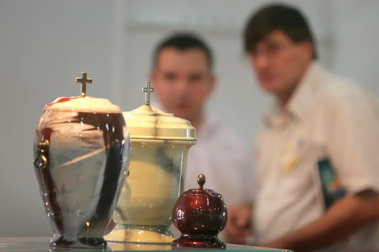 Trzy urny pogrzebowe z krzyżami, w tle niewyraźne postacie. Temat kremacji budzi dyskusje, nawet wśród hierarchów Kościoła, jak papież o kremacji.