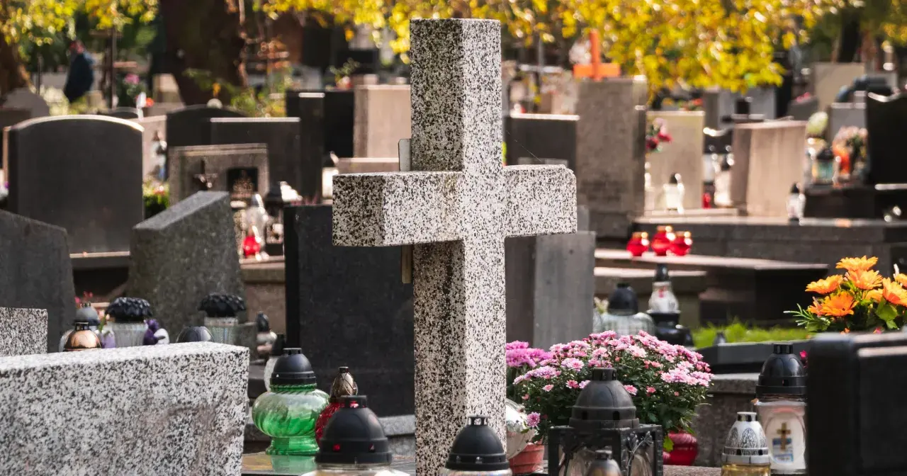 Kamienny krzyż i znicze na cmentarzu. W tle nagrobki i jesienne liście. Warto zadbać o porządek, by wiedzieć, jak usunąć chwasty na cmentarzu.