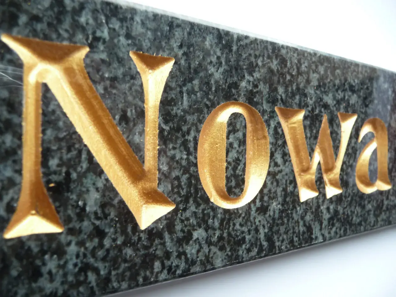Złote litery kute na pomnik, wyraz "Nowa", na ciemnym, granitowym tle.