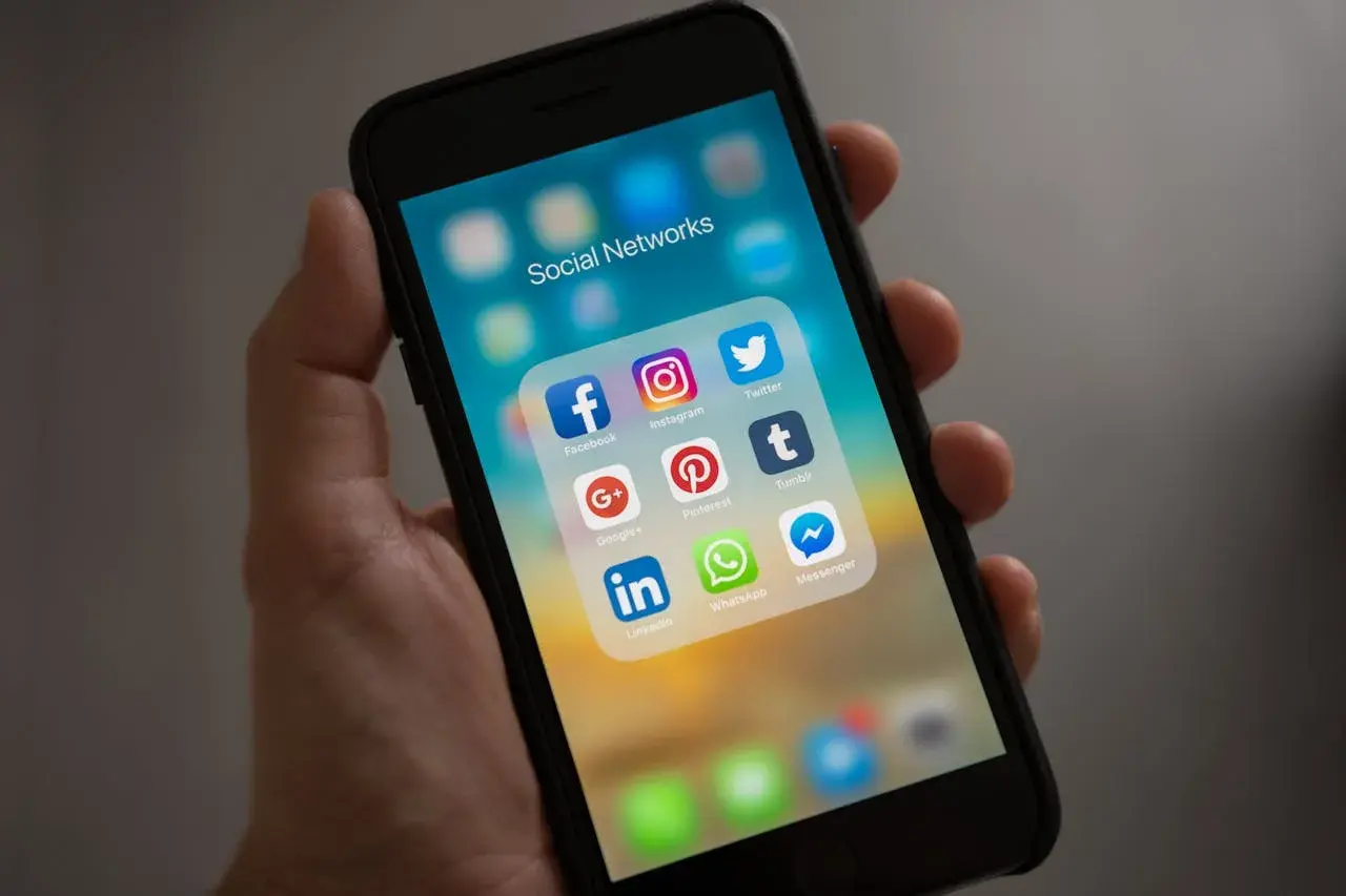 Dłoń trzyma smartfon z ikonami mediów społecznościowych. Folder "Social Networks" zawiera Facebooka, Instagram, Twitter, Google+, Pinterest, Tumblr, LinkedIn, WhatsApp i Messenger.