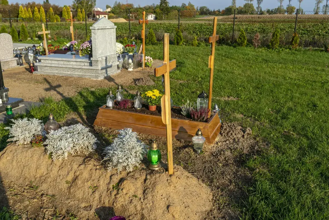 Drewniana trumna z kwiatami i krzyżami, obok kamienny nagrobek. Tak wygląda grób, ozdobiony zniczami i zielenią.