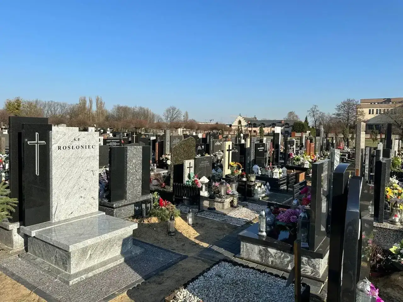 Cmentarz z nagrobkami, krzyżami i kwiatami. W oddali budynki. Może to miejsce po ekshumacji.