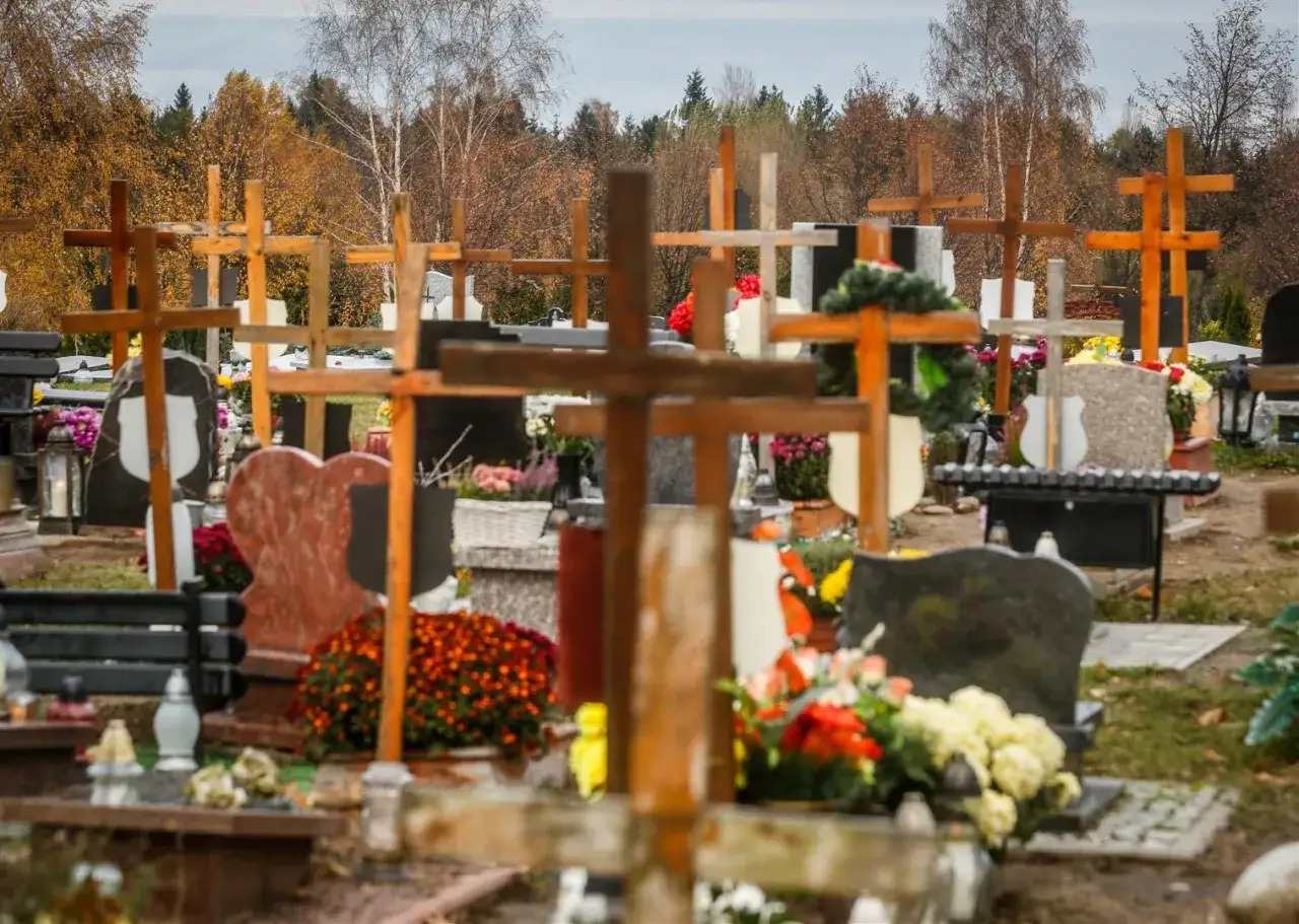 Cmentarz jesienią, wiele drewnianych krzyży i nagrobków, ozdobionych kwiatami. W tle jesienne drzewa.