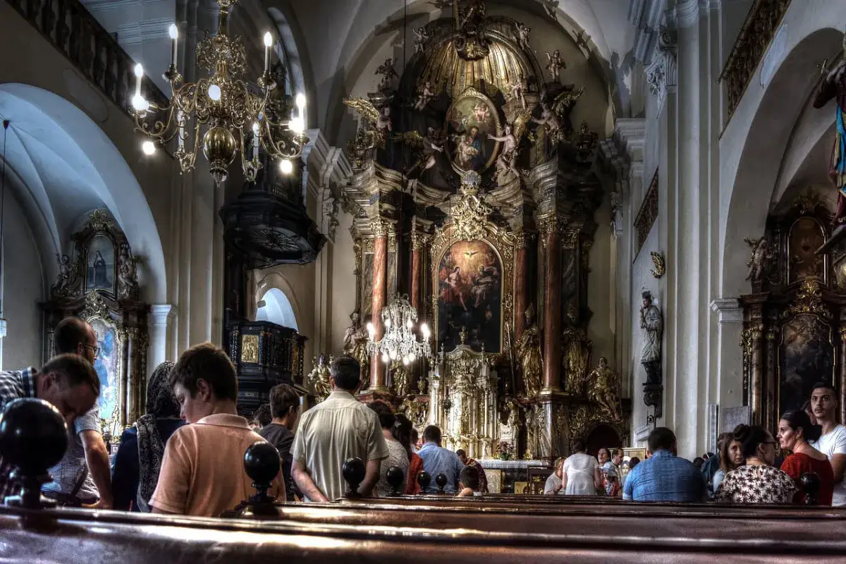Wnętrze kościoła z wiernymi siedzącymi w ławkach. Złoty ołtarz i żyrandol. Czas mszy świętej jest różny, ale tu trwa spokojne nabożeństwo.