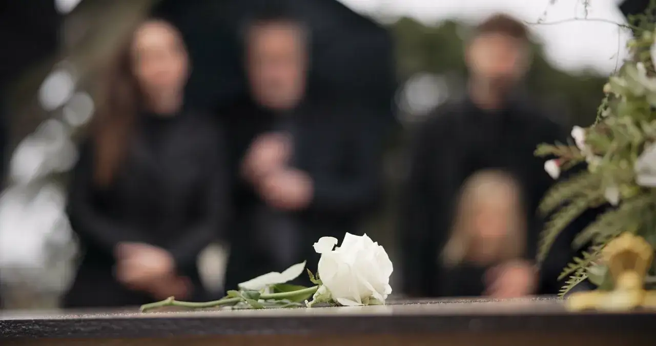 Biała róża na kamieniu, symbol żałoby. Ludzie w czerni, jak nie płakać na pogrzebie, wspominają bliską osobę.