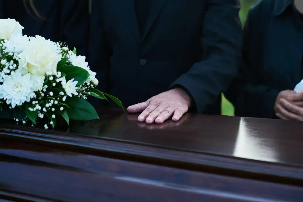 Dłoń spoczywa na ciemnej trumnie obok białych kwiatów. Uczestnicy w czerni żegnają bliską osobę podczas pogrzebu świeckiego.