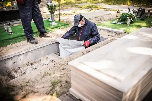Ekshumacja zwłok po 40 latach. Pracownik w masce i rękawiczkach wyciąga worek z grobu. Obok leży nowa trumna.
