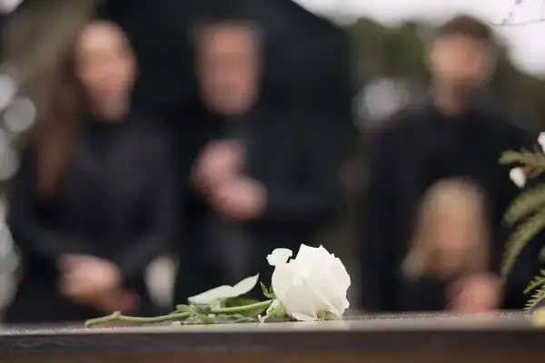 Biała róża na kamiennej płycie, w tle zamazane sylwetki żałobników. Czas organizacji pogrzebu jest trudny.